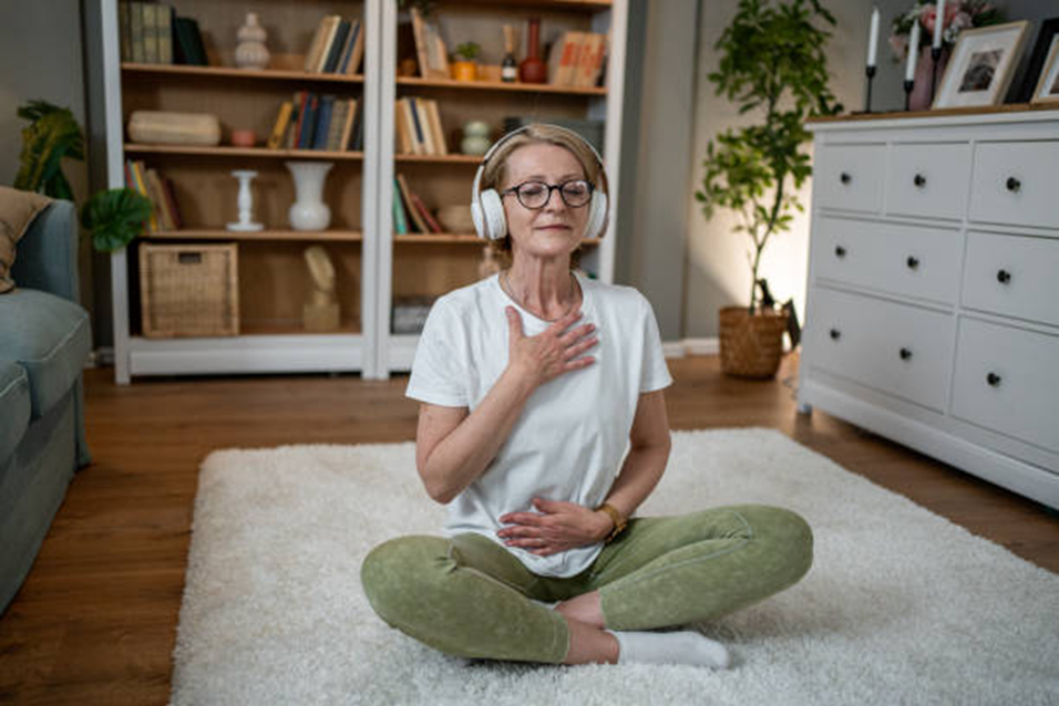 Older woman in headphones sitting cross-legged at home, hand on heart, paying attention
