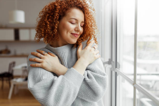 Curly-haired woman hugging herself by a window, practicing gentle self-love